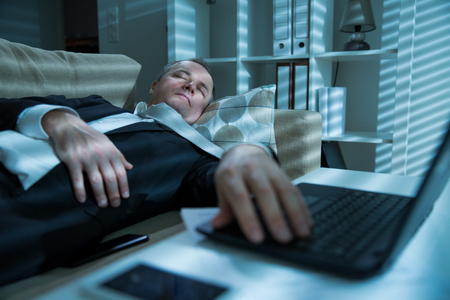Businessman in suit lying on a couch with two cellphones and laptops, sleeping. Exhausted man relaxing in office late at night. Responsible executive working fell asleep.の写真素材