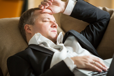 Businessman in suit lying on a couch with two cellphones and laptops, sleeping. Exhausted man relaxing in office early morning. Responsible executive working fell asleep.の写真素材