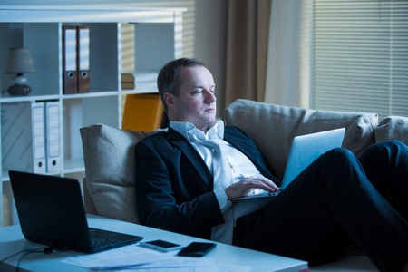 Businessman in suit lying on a couch with two cellphones and laptops. Exhausted man relaxing in office late at night. Responsible executive workingの写真素材