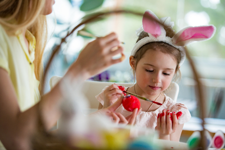 A mother and daughter celebrating Easter, painting eggs with brush. Happy family smiling and laughing. Cute little girl in bunny ears preparing the holiday.の写真素材