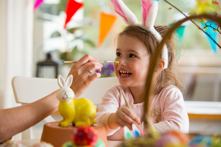 A mother and daughter celebrating Easter, painting bunny with brush in yellow color. Happy family smiling and laughing. Cute little girl in bunny ears preparing the holiday.の写真素材
