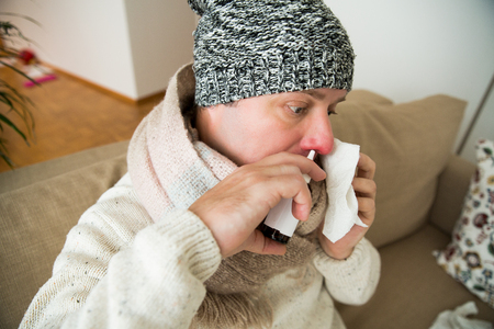 Sick man catch cold. Ill person sneezing, coughing, got flu, having red runny nose, spraying medication. Sitting at home on couch.の写真素材