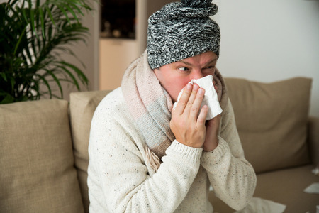 Person with cold blowing his nose. Sick man wearing scarf sitting on couch at home. Man with red running nose and cough.の写真素材