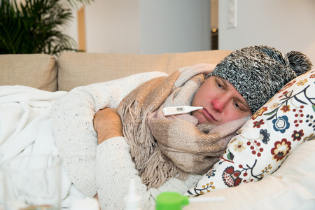 Sick man wearing scarf lying on couch at home under a blanket checking temperature. Living room with table full of medicine and pills. Man with running nose and cough.の写真素材