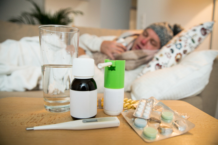Sick man wearing scarf lying on couch at home under a blanket checking temperature. Living room with table full of medicine and pills. Man with running nose and cough.の写真素材