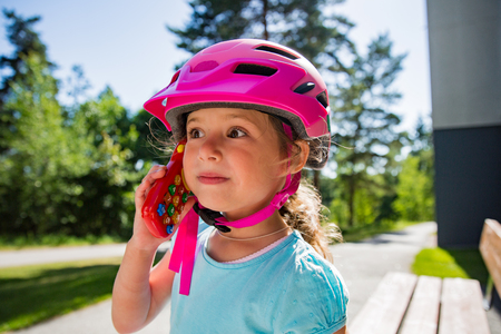 Little girl in helmet sitting on bench playing with toy cell phone. Sunny summer day in suburb. Happy childhood.の写真素材