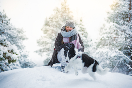 Woman playing with dog in snowy forest, enjoying the weather. Running and jumping happy pet, girl laughing, having fun. Beautiful winter landscape with trees in snow.の写真素材
