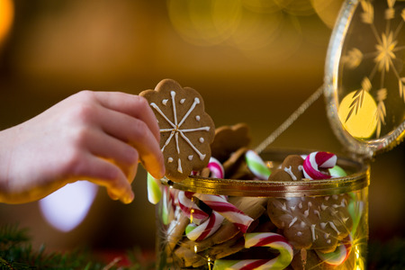 Cute little girl taking candies and gingerbread from glass jar and eating. Happy family holiday. Close-up hand. Living room decorated with lights and candles and Christmas tree. Holiday moodの写真素材