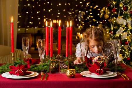 Cute little girl celebrating Christmas, taking candies and gingerbread from glass jar and eating. Served table with candles, Red tablecloth and napkins, white china, gold cutlery, crystal glasses.の写真素材