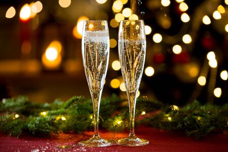 Background of two glasses of champagne, spruce branches on red table cloth. Living room decorated with lights and candles and Christmas treeの写真素材