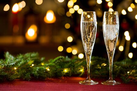 Background of two glasses of champagne, spruce branches on red table cloth. Living room decorated with lights and candles and Christmas treeの写真素材