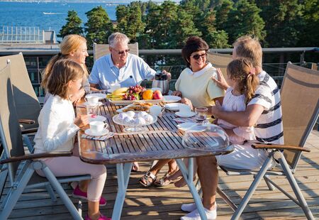 Big happy family having breakfast outdoors on terrace together, sitting around table, drinking coffee. Beautiful sea view, warm summer morning. Family bonds conceptの写真素材
