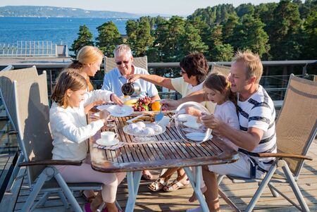 Big happy family having breakfast outdoors on terrace together, sitting around table, drinking coffee. Beautiful sea view, warm summer morning. Family bonds conceptの写真素材