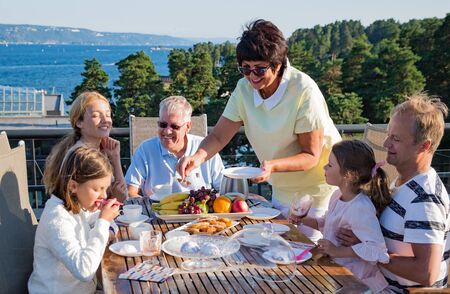 Big happy family having breakfast outdoors on terrace together, sitting around table, drinking coffee. Beautiful sea view, warm summer morning. Family bonds conceptの写真素材