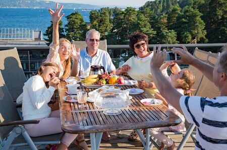 Big happy family having breakfast outdoors on terrace together, sitting around table, drinking coffee. Beautiful sea view, warm summer morning. Family bonds conceptの写真素材
