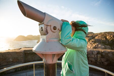 Teenage girl stands on rocky northern seashore, looking through touristic telescope, exploring the coastal rocks. Travel and enjoy adventure in Norway. Beautiful view of fjord and mountains in sunsetの写真素材
