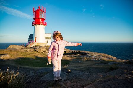 Little girl stands on rocky northern seashore, exploring the coastal rocks. Travel and enjoy a great adventure in Norway. Beautiful view of lighthouse and sea in sunset.の写真素材