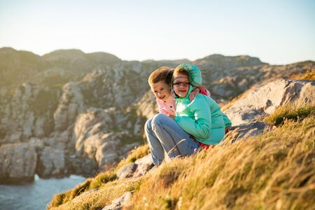 Two little girls play on rocky northern seashore. Sit, laugh, hug, explore the coastal rocks. Travel and enjoy a great adventure in Norway. Beautiful view of fjord and mountains in sunset.の写真素材