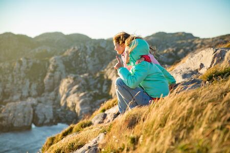 Two little girls play on rocky northern seashore. Sit, laugh, hug, explore the coastal rocks. Travel and enjoy a great adventure in Norway. Beautiful view of fjord and mountains in sunset.の写真素材