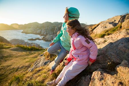 Two little girls play on rocky northern seashore. Sit, laugh, hug, explore the coastal rocks. Travel and enjoy a great adventure in Norway. Beautiful view of fjord and mountains in sunset.の写真素材