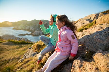 Two little girls play on rocky northern seashore. Sit, laugh, hug, explore the coastal rocks. Travel and enjoy a great adventure in Norway. Beautiful view of fjord and mountains in sunset.の写真素材