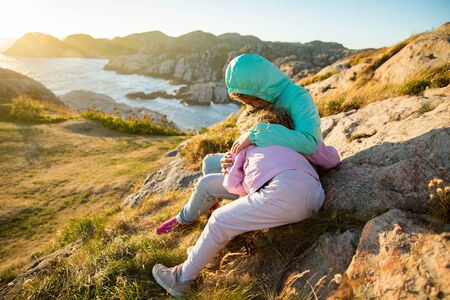 Two little girls play on rocky northern seashore. Sit, laugh, hug, explore the coastal rocks. Travel and enjoy a great adventure in Norway. Beautiful view of fjord and mountains in sunset.の写真素材