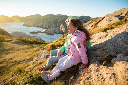 Two little girls play on rocky northern seashore. Sit, laugh, hug, explore the coastal rocks. Travel and enjoy a great adventure in Norway. Beautiful view of fjord and mountains in sunset.の写真素材