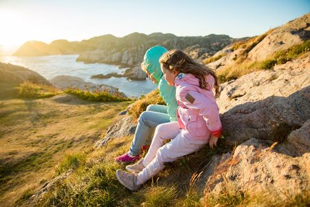 Two little girls play on rocky northern seashore. Sit, laugh, hug, explore the coastal rocks. Travel and enjoy a great adventure in Norway. Beautiful view of fjord and mountains in sunset.の写真素材