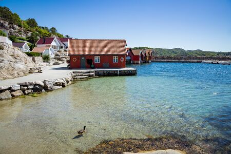 Lindesnes, Norway - July 7, 2018:Serene scandinavian summer landscape of countryside on south coast of Norway. Sunny sand beach with turquoise quiet water. Boats and houses with Norwegian flag.の写真素材