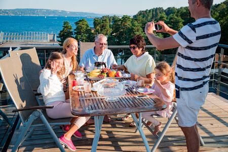 Big happy family having breakfast outdoors on terrace together, sitting around table, drinking coffee. Father taking selfie on phone. Beautiful sea view, warm summer morning. Family portrait.の写真素材