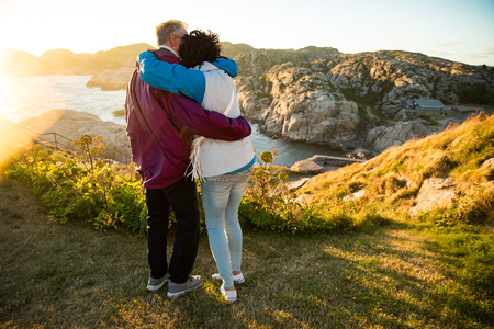 Loving mature couple traveling, standing on the top of rock, exploring. Active man and woman hugging and kissing, Happily smiling. Scenic view of mountains and sea on sunset. Norway, Lindesnesの写真素材