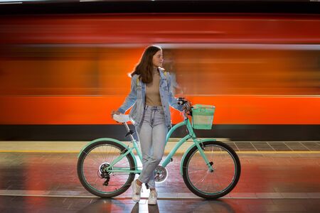 Teenager girl in jeans with yellow backpack and bike standing on metro station, waiting for train, smiling, laughing. Orange train passing by behind the girl. Futuristic subway station. Finland, Espooの写真素材