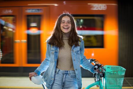 Teenager girl in jeans with yellow backpack and bike standing on metro station, waiting for train, smiling, laughing. Orange train passing by behind the girl. Futuristic subway station. Finland, Espooの写真素材
