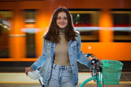 Teenager girl in jeans with yellow backpack and bike standing on metro station, waiting for train, smiling, laughing. Orange train passing by behind the girl. Futuristic subway station. Finland, Espooの写真素材