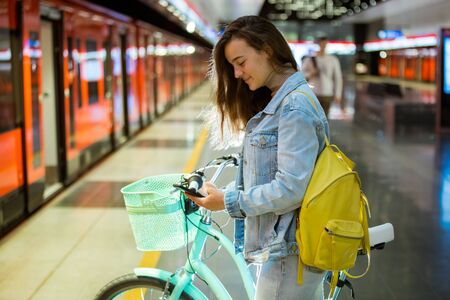 Teenager girl with backpack and bike standing on metro station holding smart phone in hand, scrolling and texting, smiling and laughing. Futuristic bright subway station. Finland, Espooの写真素材