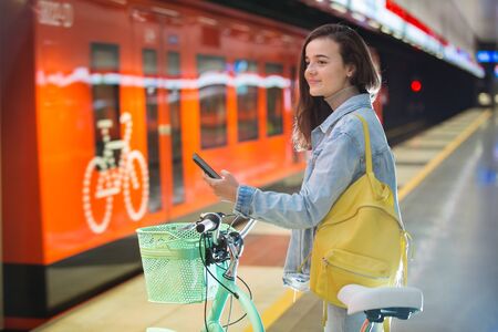Teenager girl with backpack and bike standing on metro station holding smart phone in hand, scrolling and texting, smiling and laughing. Futuristic bright subway station. Finland, Espooの写真素材