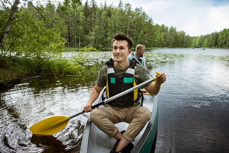 Two men in life vests canoeing in forest lake. Water surface covered with water lilies. Tourists traveling in Finland, having adventure.の写真素材