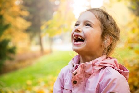 Cute little girl with missing teeth playing with yellow fallen leaves in autumn forest. Happy child laughing and smiling. Sunny autumn forest, sun beam.の写真素材