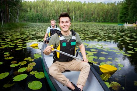 Two men in life vests canoeing in forest lake. Water surface covered with water lilies. Tourists traveling in Finland, having adventure.の写真素材