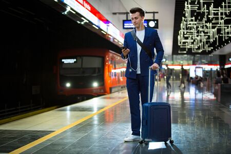 Young stylish handsome man in suit with suitcase standing on metro station holding smart phone in hand, scrolling and texting, smiling and laughing.  Orange train passing by. Futuristic bright subwayの写真素材