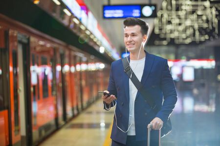 Young stylish handsome man in suit with suitcase standing on metro station holding smart phone in hand, scrolling and texting, smiling and laughing.  Orange train passing by. Futuristic bright subwayの写真素材