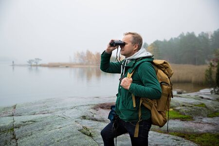 Mature man exploring Finland in the fall, looking into fog through binoculars. Hiker with big...