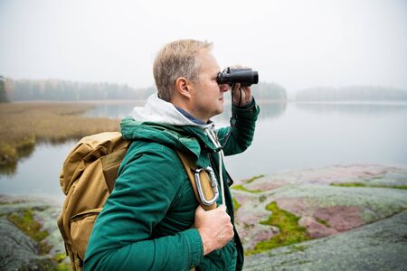 Mature man exploring Finland in the fall, looking into fog through binoculars. Hiker with big backpack standing on mossy rock. Scandinavian landscape with misty sea and autumn forest.の写真素材
