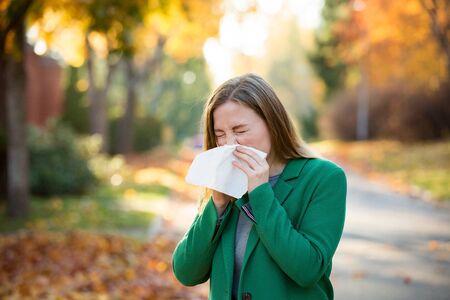 Sick young woman with cold and flu standing outdoors, sneezing, wiping nose with handkerchief, coughing. Autumn street backgroundの写真素材