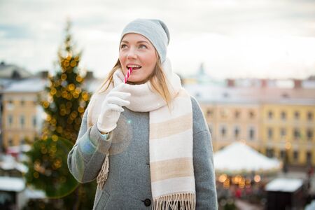 Young woman in Christmas market eating candy cane wearing knitted warm hat and scarf. Illuminated and decorated fair kiosks, shops and carousel on background. Helsinki, Finlandの写真素材