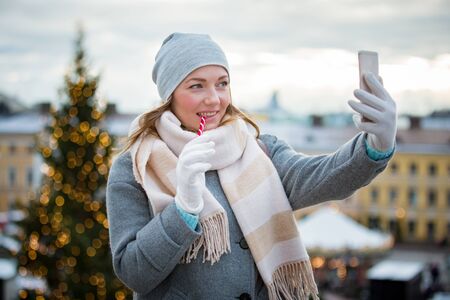 Young woman in Christmas market eating candy cane and taking selfie. Girl in knitted warm hat and scarf. Illuminated and decorated fair kiosks, shops and carousel on background. Helsinki, Finlandの写真素材