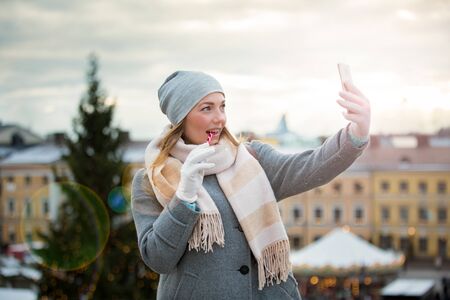 Young woman in Christmas market eating candy cane and taking selfie. Girl in knitted warm hat and scarf. Illuminated and decorated fair kiosks, shops and carousel on background. Helsinki, Finlandの写真素材