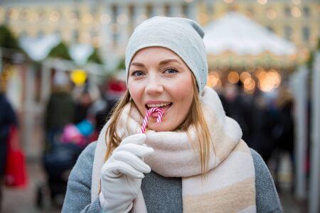 Young woman in Christmas market eating candy cane wearing knitted warm hat and scarf. Illuminated and decorated fair kiosks, shops and carousel on background. Helsinki, Finlandの写真素材