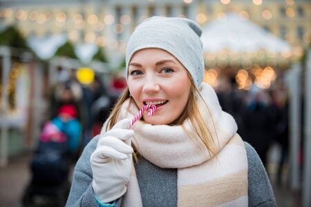 Young woman in Christmas market eating candy cane wearing knitted warm hat and scarf. Illuminated and decorated fair kiosks, shops and carousel on background. Helsinki, Finlandの写真素材
