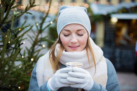 Young woman in Christmas market drinking cup of hot chocolate with marshmallow wearing knitted warm hat and scarf. Illuminated and decorated fair kiosks and shops on background. Helsinki, Finlandの写真素材